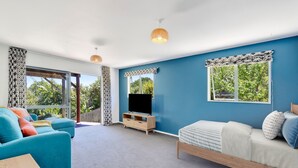 A room with a blue wall, a bed, a sofa, a TV stand, and a window with a view of greenery.