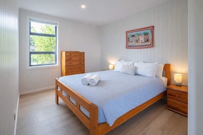 A bedroom with a wooden bed, a dresser, a nightstand, a window with a view of greenery, and a painting on the wall.