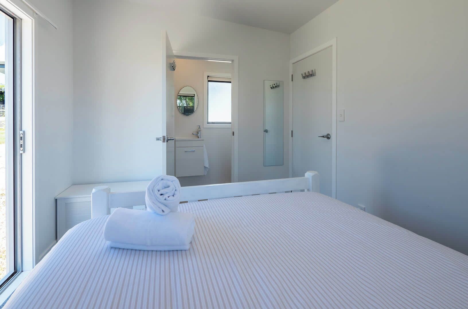A bedroom with a large bed, a window, a bathroom with a mirror, and a door.