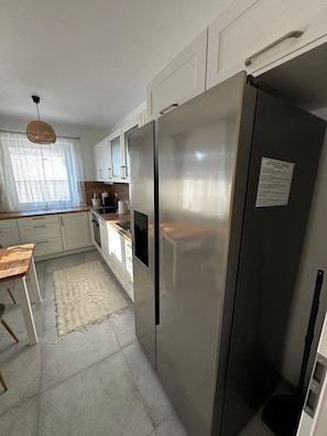 A compact kitchen with white cabinets, a stainless steel refrigerator, a wooden dining table, and a woven pendant light.