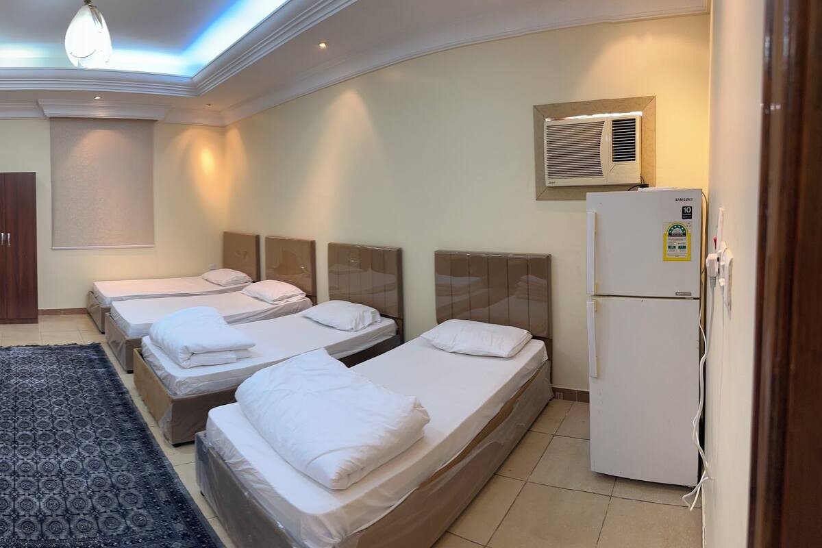 Habitación de hotel con tres camas, un sofá rojo, una alfombra estampada, una lámpara de techo y un aire acondicionado instalado en la pared.