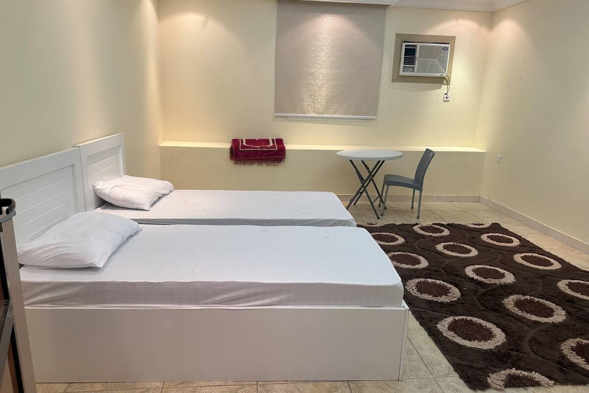Una habitación con dos camas, una mesita, una silla, una ventana con persianas y un equipo de aire acondicionado.
