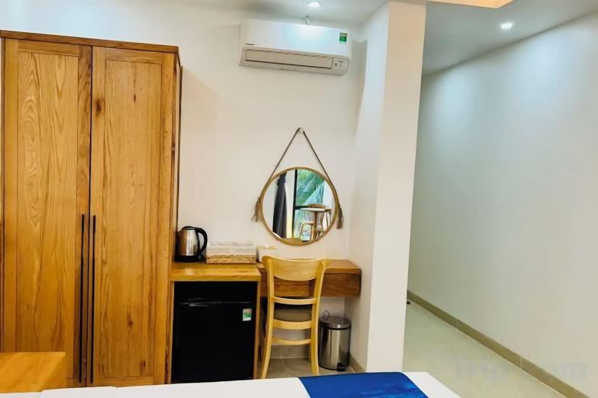 Une chambre d’hôtel comprenant un lit, une armoire en bois, un bureau avec une chaise, un petit réfrigérateur et un climatiseur fixé au mur.