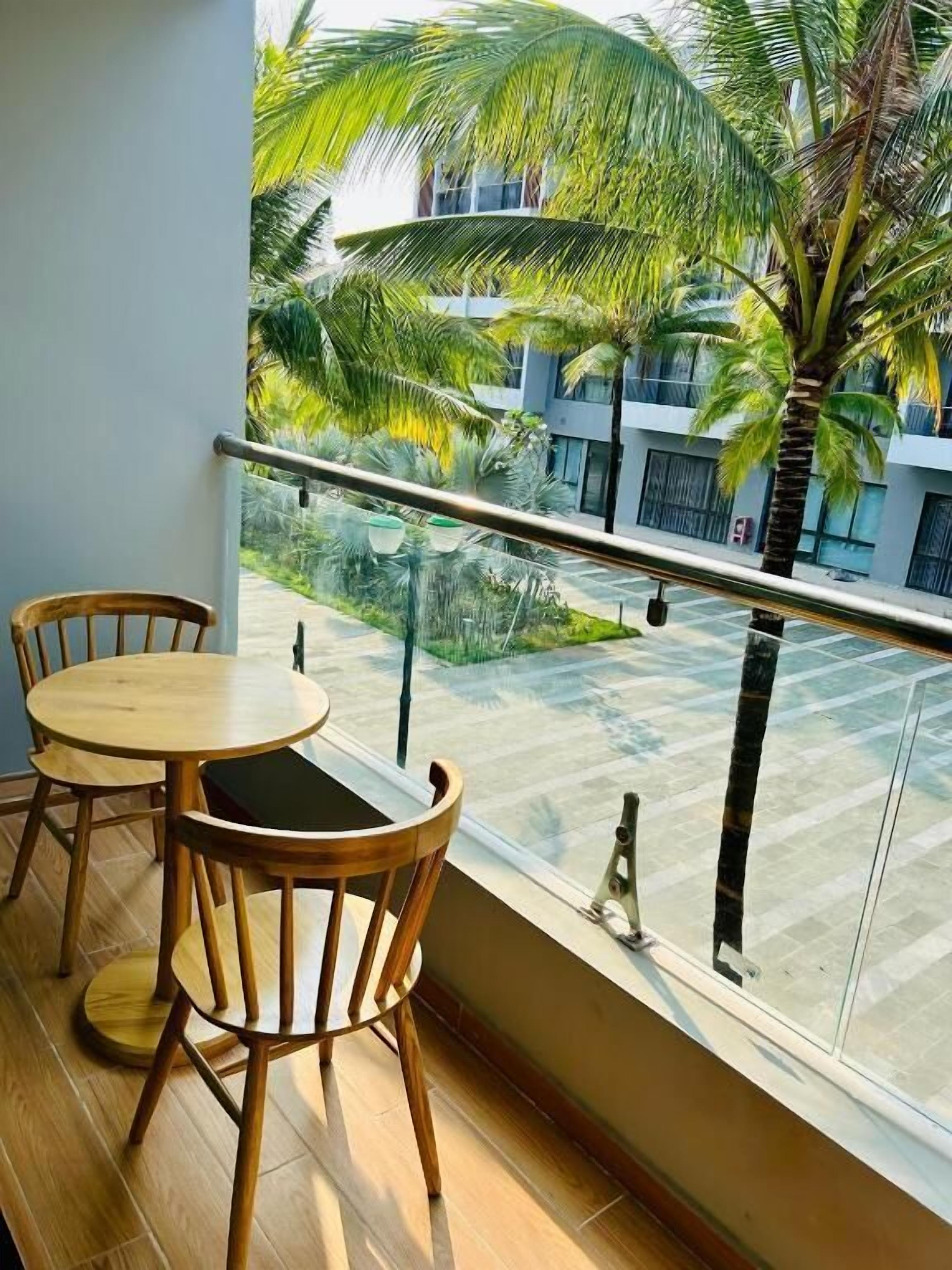 Varanda com duas cadeiras de madeira e uma mesa pequena, com vista para um pátio com palmeiras e um prédio ao fundo.