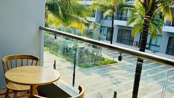 Varanda com duas cadeiras de madeira e uma mesa pequena, com vista para um pátio com palmeiras e um prédio ao fundo.