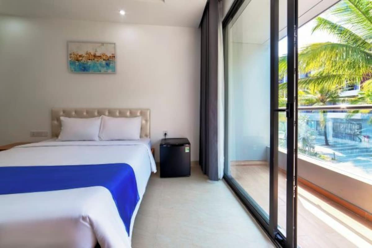 A hotel room with a bed, a small black luggage storage unit, and a large window with a view of palm trees and a pool.