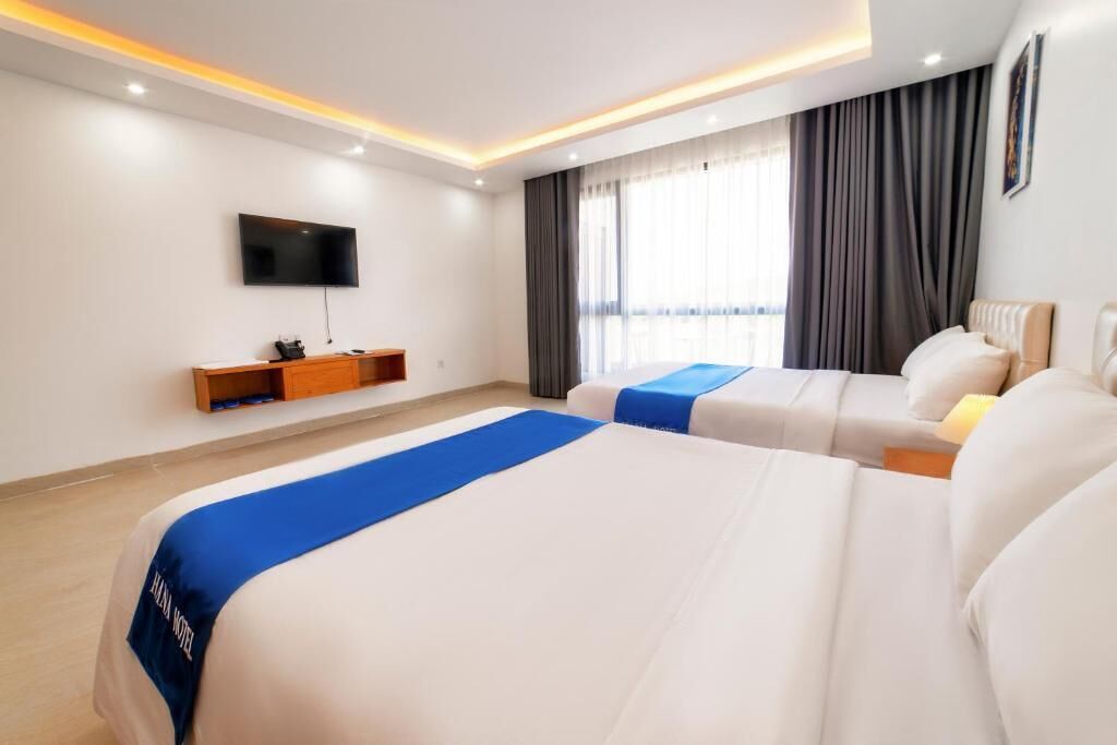 A hotel room with two beds, a TV mounted on the wall, a window with curtains, and a bedside table.