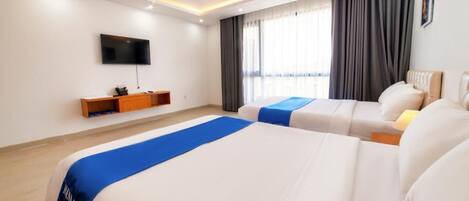 A hotel room with two beds, a TV mounted on the wall, a window with curtains, and a bedside table.