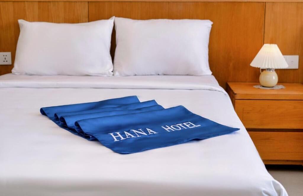 A neatly made bed with white linens and a blue towel with "HANA HOTEL" text.