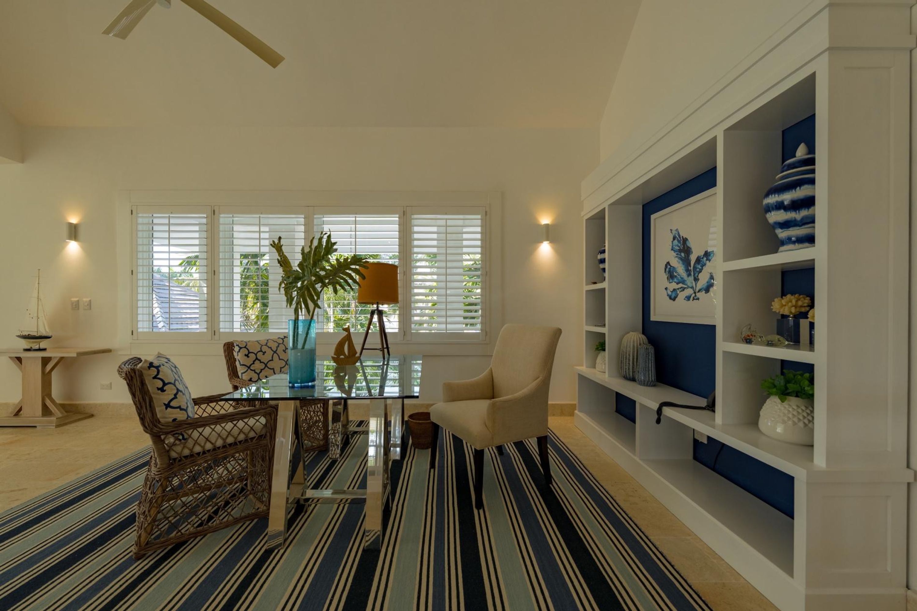 Un salon avec un tapis à rayures, une table en verre, des chaises en osier, un vase bleu et un fauteuil blanc.