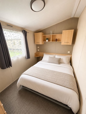 A bedroom with a bed, a window with curtains, a shelf with decorative items, and a wall-mounted light fixture. - Singh&Leite Family L6 (Clacton-on-Sea)