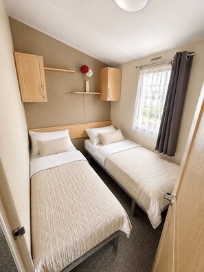 Ein kleines Zimmer mit zwei Betten, einem kleinen Regal mit Blumen und einem Fenster mit Vorhängen.
