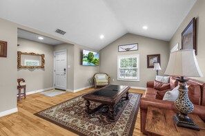 A spacious living room with hardwood flooring, a large coffee table, a leather sofa, and a television mounted on the wall.