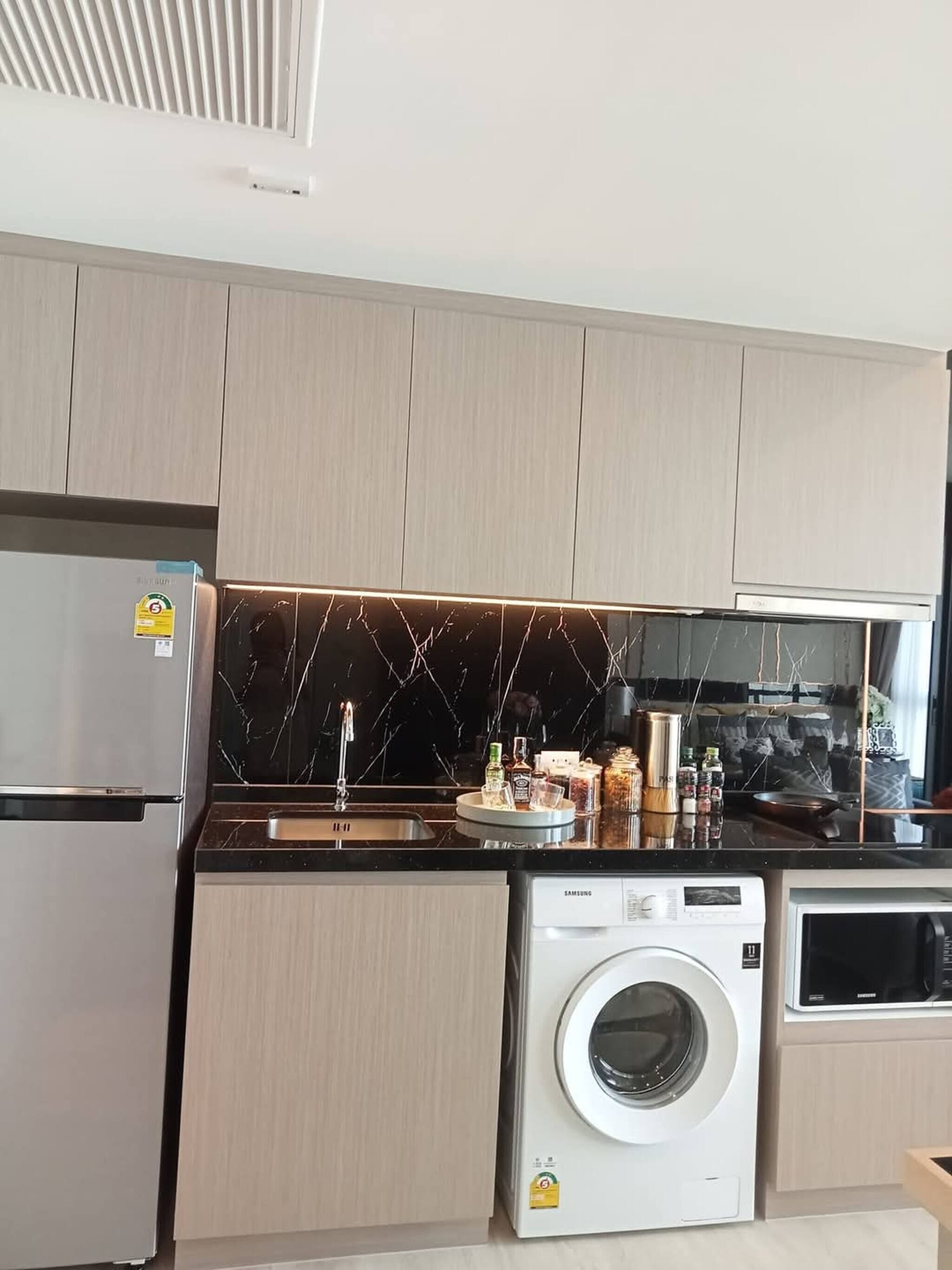 A modern kitchen with light wood cabinets, a stainless steel refrigerator, a built-in oven, a microwave, and a washing machine.