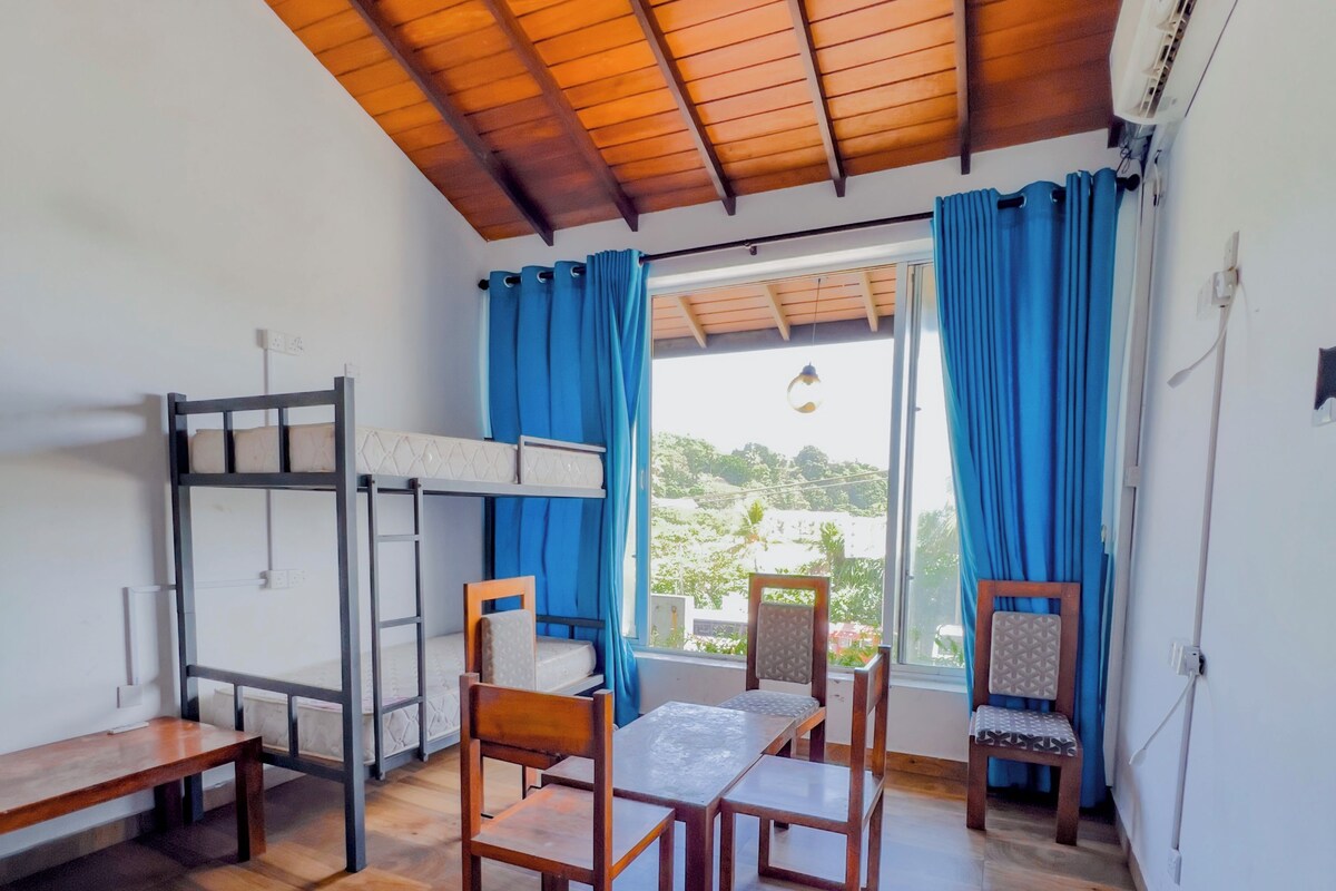 Habitación con literas, una mesa, sillas y vista a través de una ventana con cortinas.