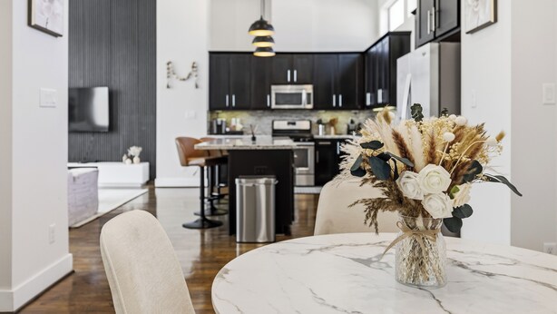 A modern kitchen with dark cabinets, a central island, and a dining area with a marble table and chairs. - Luxury 3Bed/3.5Bath Townhome 10 Minutes from Downtown HTX (Houston)