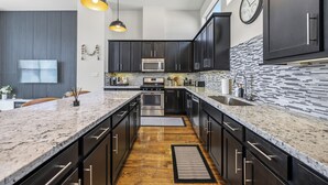 A modern kitchen with dark cabinetry, a central island, stainless steel appliances, and a patterned backsplash. - Luxury 3Bed/3.5Bath Townhome 10 Minutes from Downtown HTX (Houston)