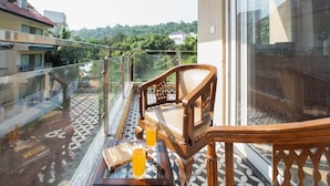 Un balcon avec une chaise en bois, une table en verre sur laquelle se trouvent un livre et des lunettes, et une vue sur un paysage verdoyant.