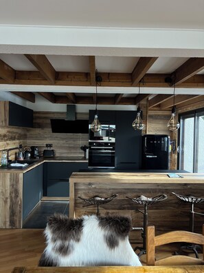 A modern kitchen with wooden beams, a black refrigerator, and a dining area with a wooden table and chairs. - Pilgrim's Cabin - A breathtaking view and close to everything! (Font-Romeu-Odeillo-Via)