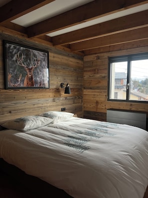 A bedroom with a wooden ceiling, a bed with white bedding, a framed deer picture on the wall, and a window with a view of buildings. - Pilgrim's Cabin - A breathtaking view and close to everything! (Font-Romeu-Odeillo-Via)