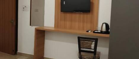 Quarto de hotel com uma cama, uma mesa de escritório com cadeira, uma televisão na parede, um guarda-roupa e uma pequena mesa com chaleira.