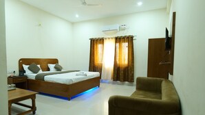 A hotel room with a bed, a wooden nightstand, a table, a chair, a TV, and an air conditioning unit. - Comfort Meets Elegance Orchha ,“Your Perfect Stay, Every Time (Orchha)