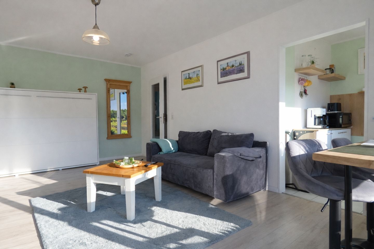 Uma sala de estar moderna com um sofá cinzento, uma mesa de centro de madeira e uma mesa de jantar com cadeiras. Há quadros nas paredes e uma área de cozinha com micro-ondas e armários.