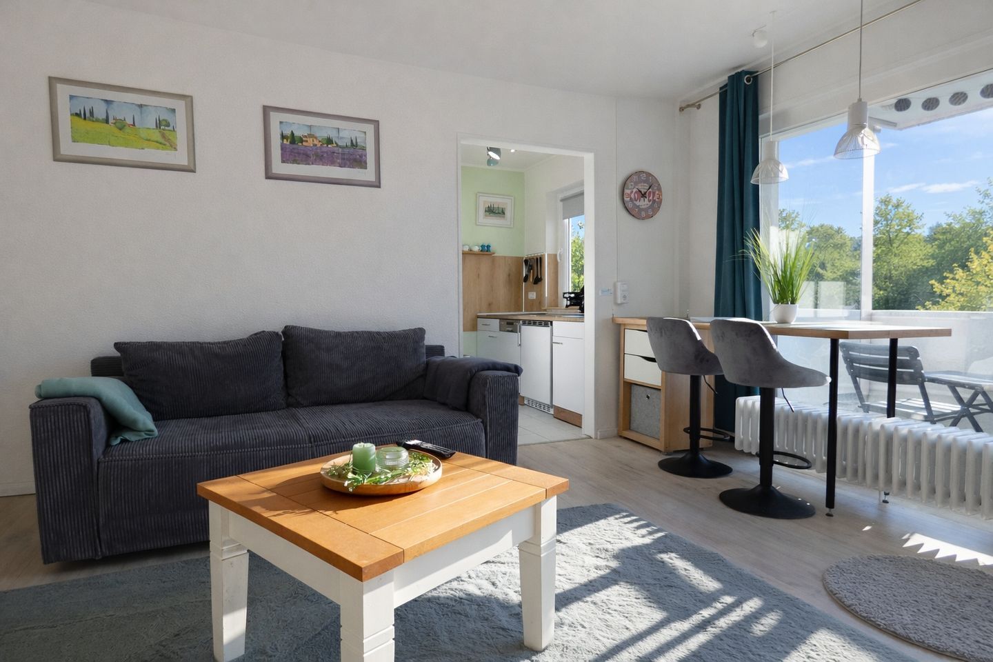 A modern living room with a grey sofa, a wooden coffee table with a green candle, and a view of a kitchen and balcony.