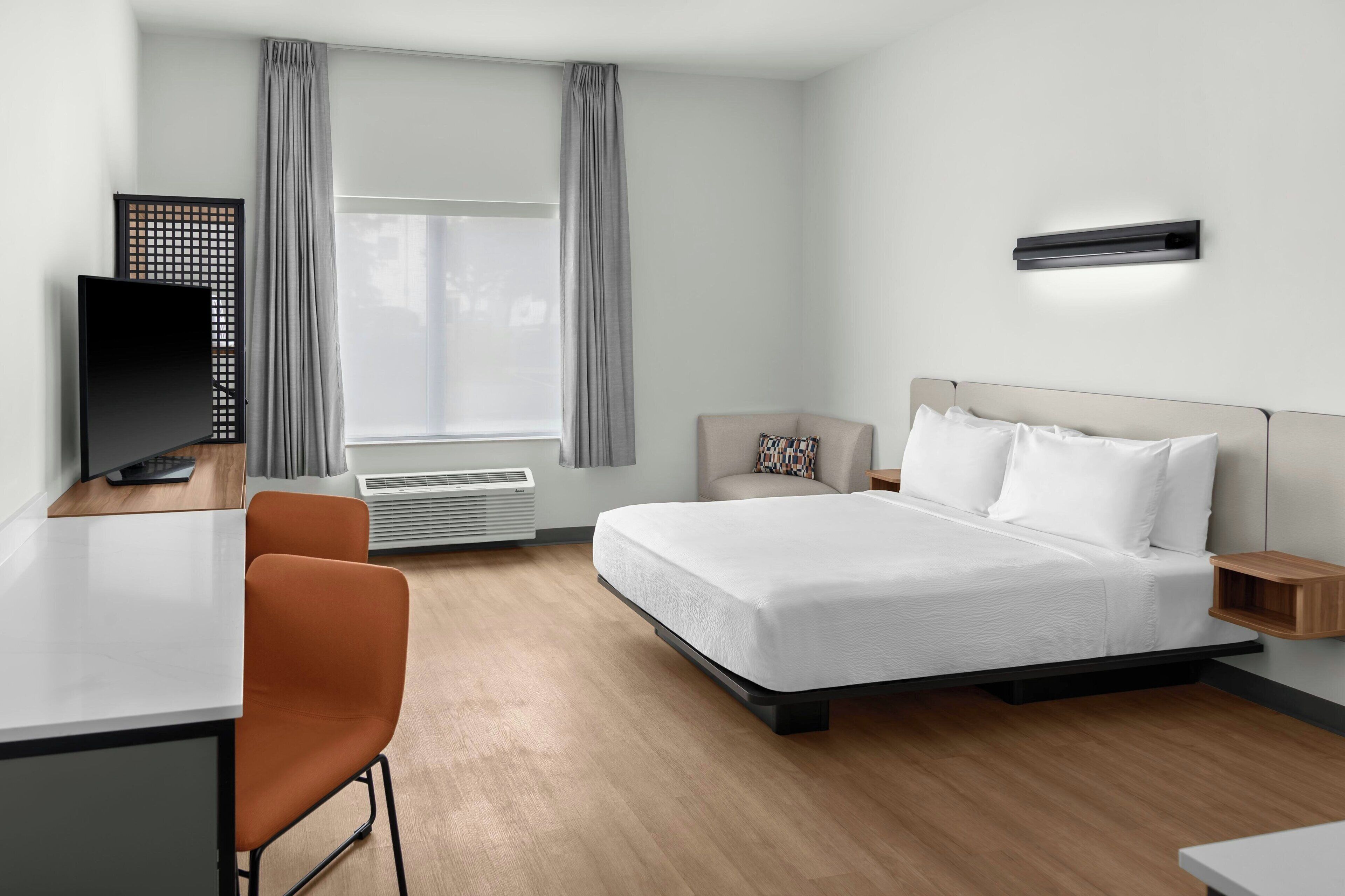 Habitación de hotel con cama, escritorio, silla, televisor y ventana con cortinas.