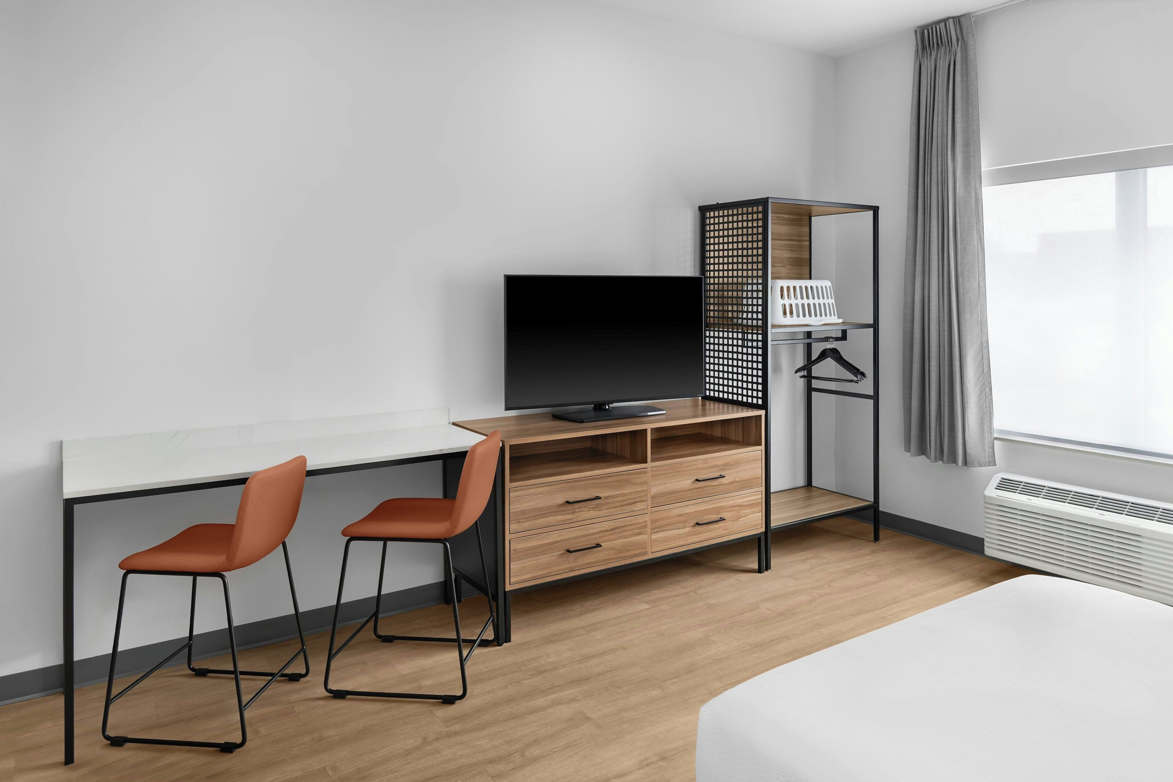 A hotel room with a bed, a desk with a TV, two chairs, a wardrobe, and a window with curtains.