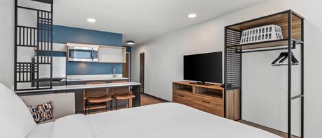 A modern hotel room with a bed, a kitchenette, a TV, and a built-in wardrobe.