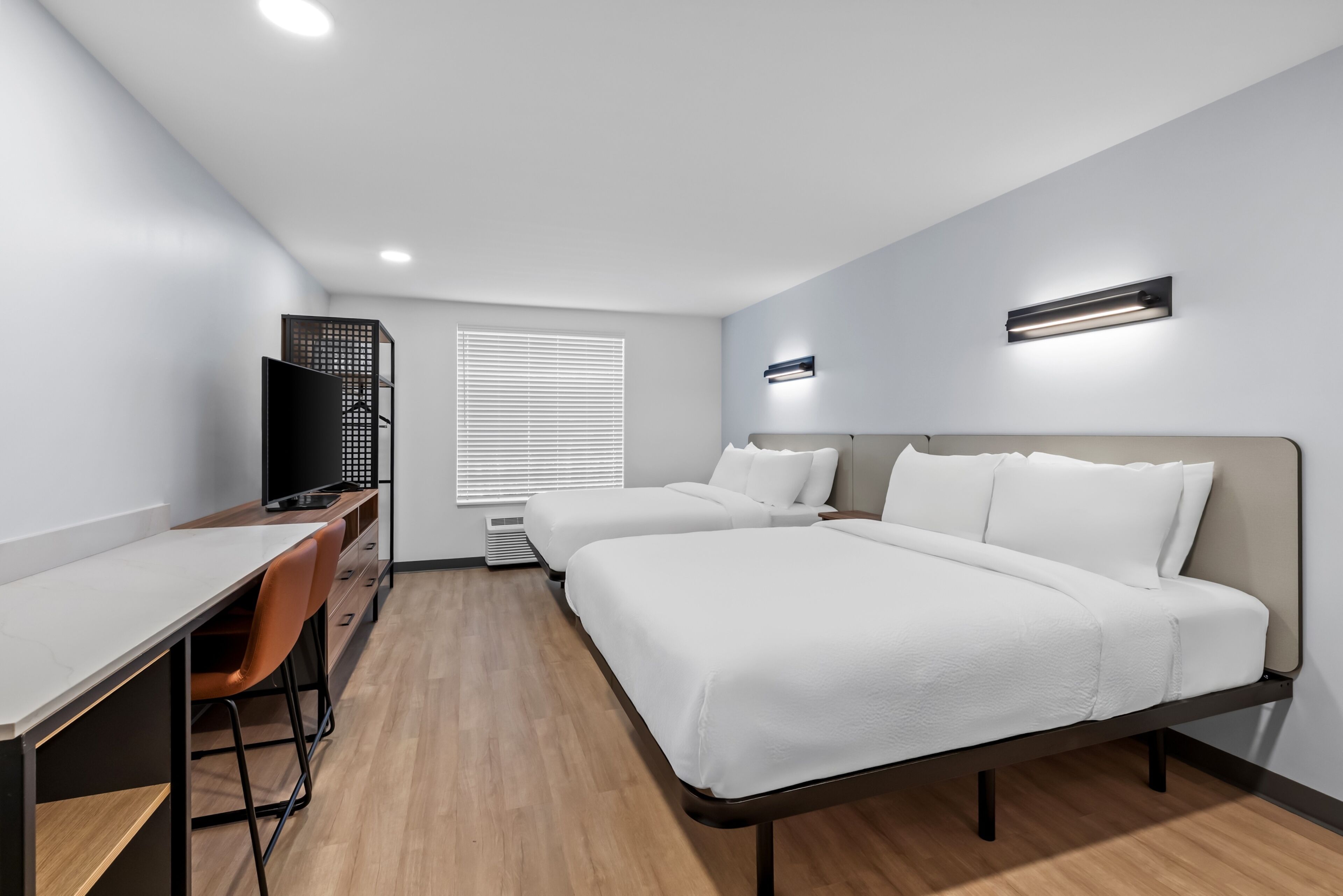 A modern hotel room with a desk, two beds, a TV, and a window with blinds.
