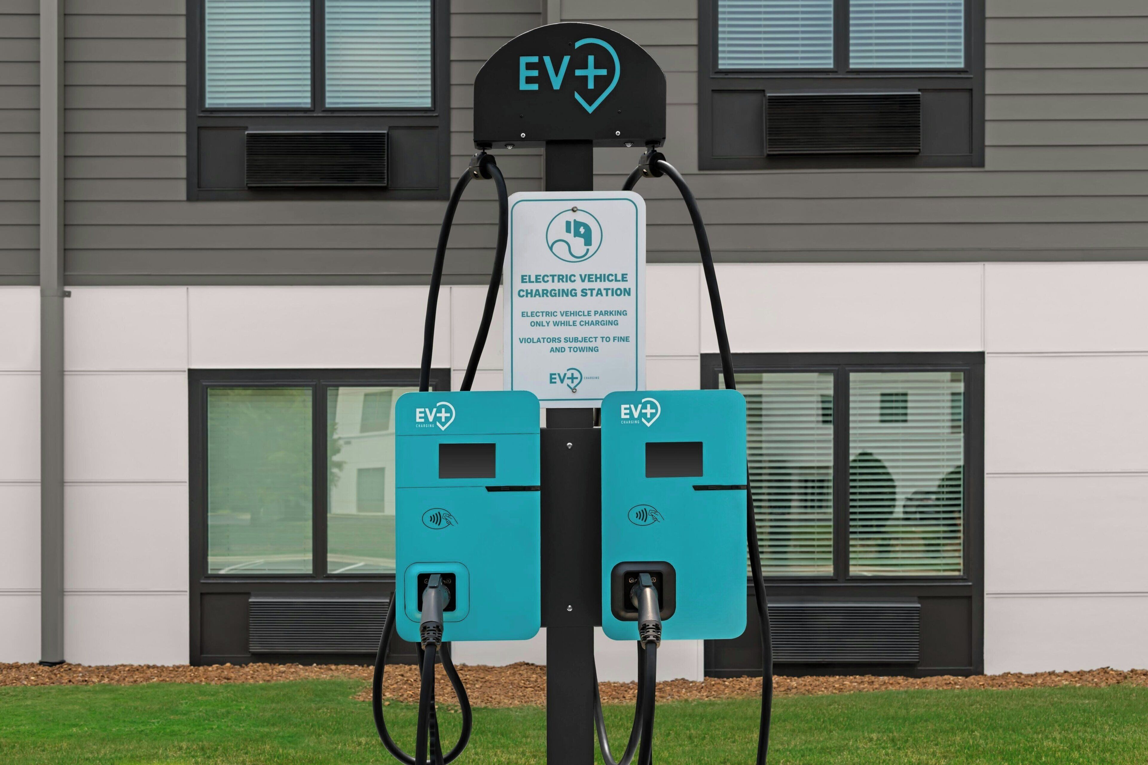 Una stazione di ricarica per veicoli elettrici con due punti di ricarica, un segnale che indica la ricarica EV+ e un edificio con ampie vetrate sullo sfondo.