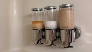 Un baño con tres dispensadores de vidrio fijos en la pared, cada uno etiquetado con un tipo diferente de jabón líquido.