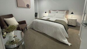 A modern hotel room with a bed, a chair, a bedside table, a lamp, and a wall art piece. - Central Albany, Popular modern 3 Beds +1 Carpark apartment (Auckland)