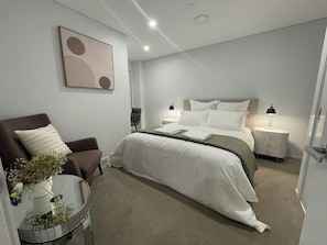 A modern hotel room with a bed, a chair, a bedside table, a lamp, and a wall art piece. - Central Albany, Popular modern 3 Beds +1 Carpark apartment (Auckland)