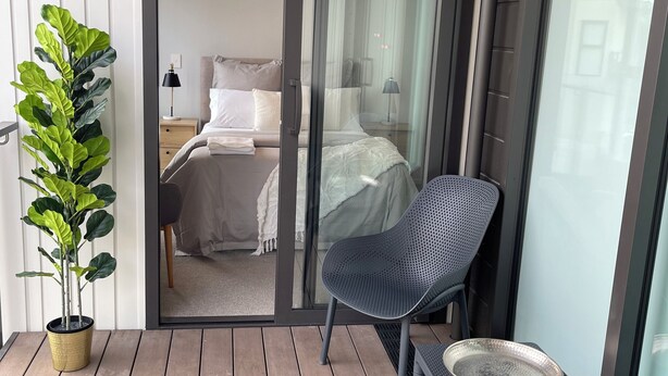 A balcony with a potted plant, two chairs, a small table, and a sliding glass door leading to a bedroom with a bed, nightstand, and lamp. - Central Albany, Popular modern 3 Beds +1 Carpark apartment (Auckland)