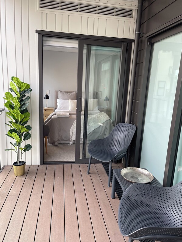 A balcony with a potted plant, two chairs, a small table, and a sliding glass door leading to a bedroom with a bed, nightstand, and lamp. - Central Albany, Popular modern 3 Beds +1 Carpark apartment (Auckland)
