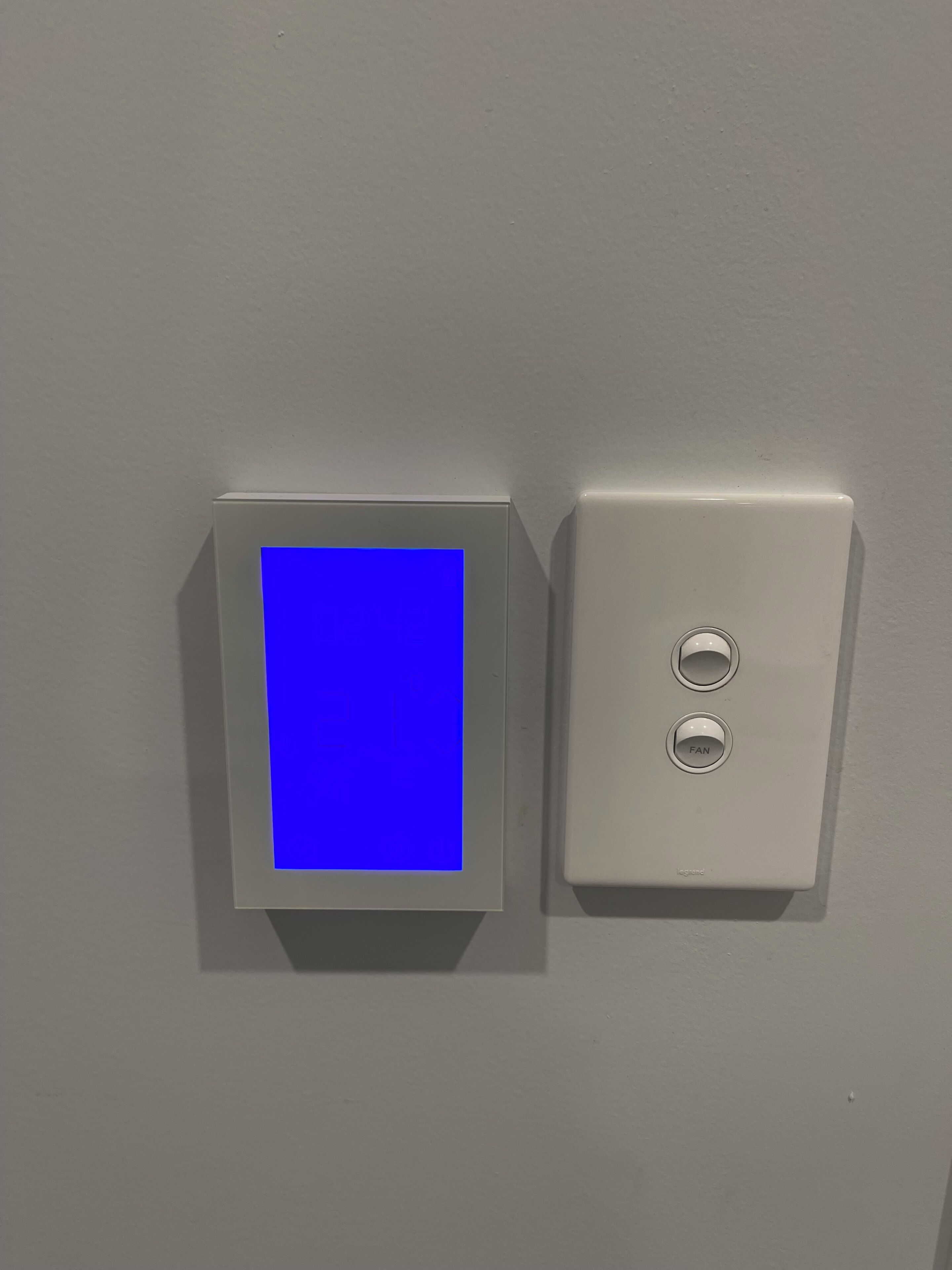 Eine wandmontierte Bedieneinheit mit blau beleuchtetem Bildschirm und zwei weißen Tasten, davon eine mit der Aufschrift 'FAN'.