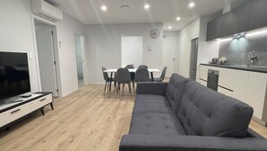 A modern living room with a grey sofa, a TV, and a dining area with chairs and a table. - Central Albany, Popular modern 3 Beds +1 Carpark apartment (Auckland)