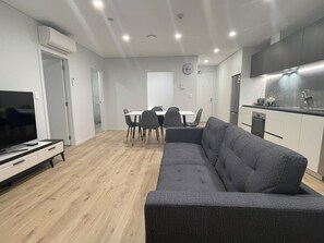 A modern living room with a grey sofa, a TV, and a dining area with chairs and a table. - Central Albany, Popular modern 3 Beds +1 Carpark apartment (Auckland)