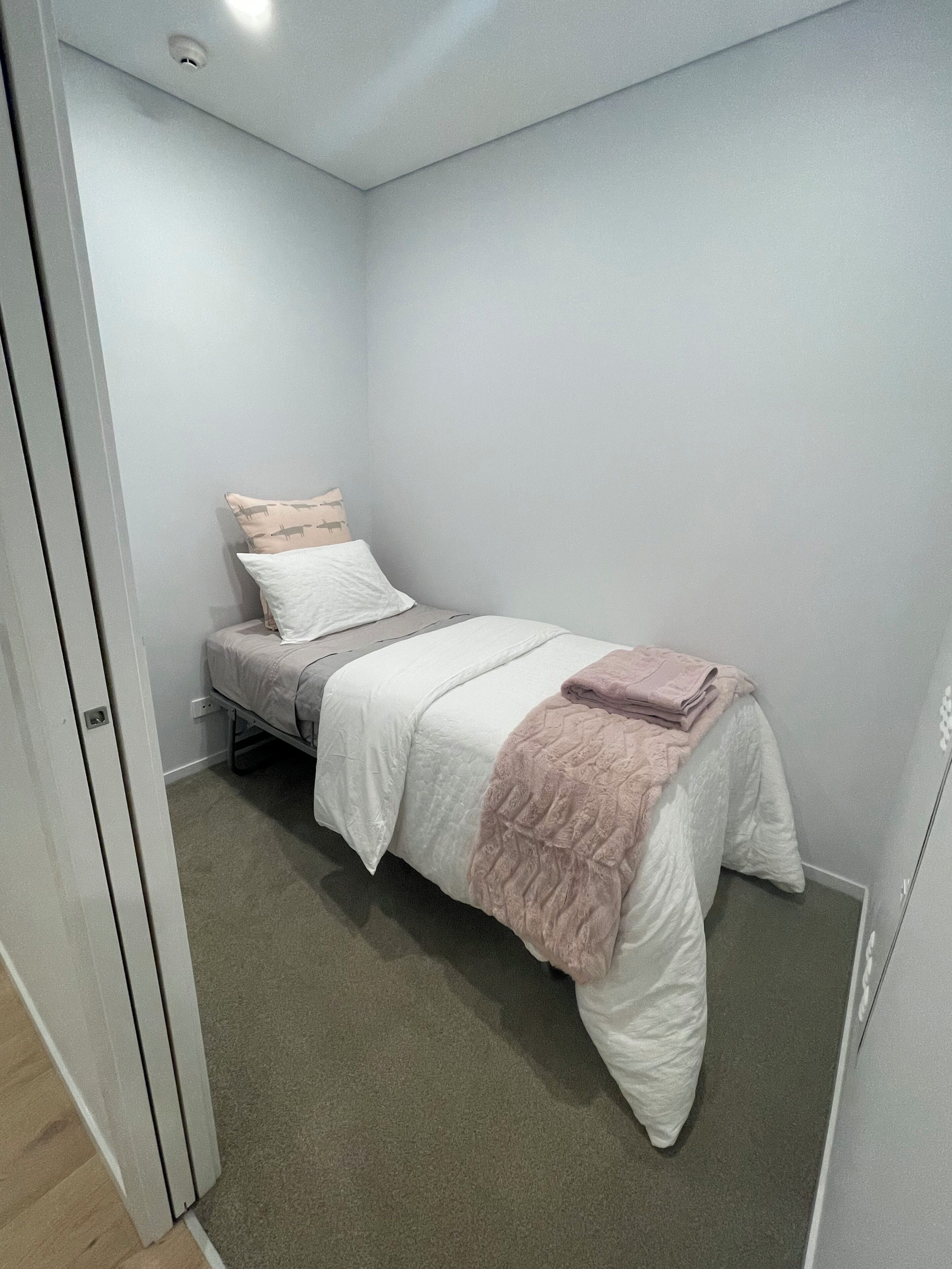 A small, single-bed room with a white wall, a door, and a carpeted floor.