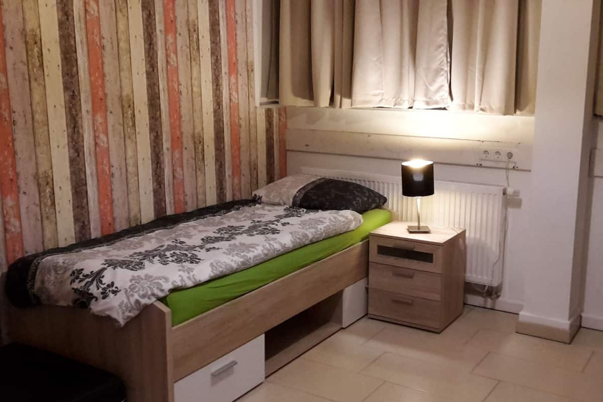 Ein kleines Zimmer mit einem Einzelbett, einem Holzkopfteil, einem Nachttisch mit Lampe und einer Heizung.