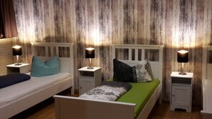 Een kamer met twee bedden, elk met een nachtkastje en een lamp. De bedden hebben beddengoed in verschillende kleuren. De muren hebben een houten structuur.