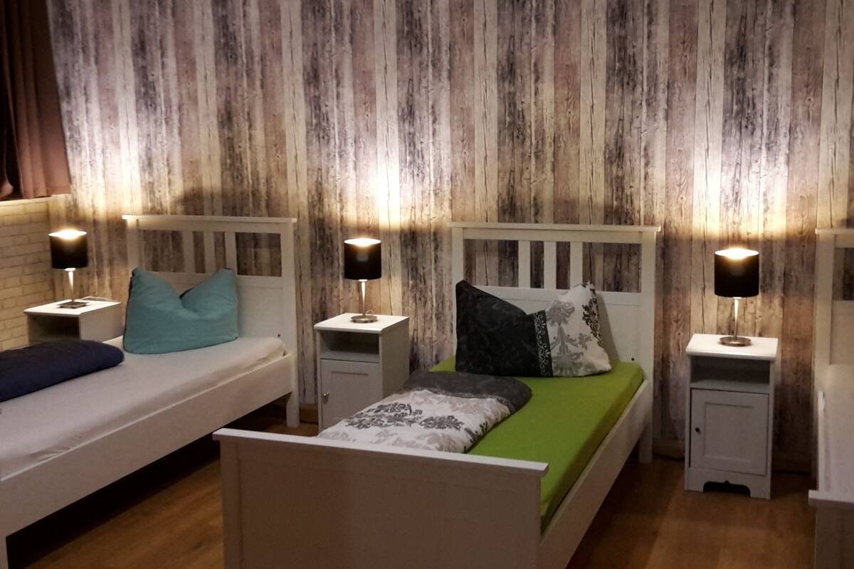 Ein Zimmer mit zwei Betten, jeweils mit einem Nachttisch und einer Lampe. Die Betten sind mit verschiedenen Bettwäschen ausgestattet. Die Wände haben eine Holzstruktur.