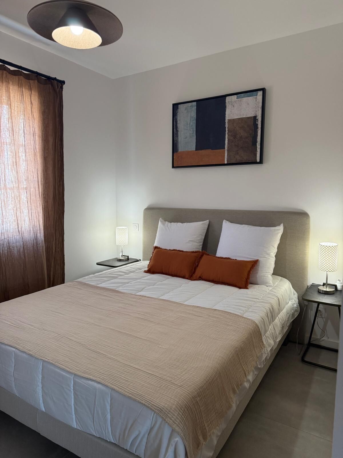 Une chambre à coucher bien rangée, avec un lit, deux tables de chevet, une œuvre d’art accrochée au mur et un luminaire au plafond.