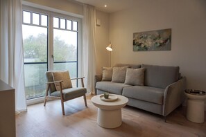 Een moderne woonkamer met een grijze bank, een houten stoel, een rond bijzettafeltje en een schilderij aan de muur.
