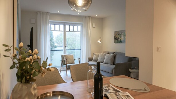 Un salon moderne avec une table à manger en bois, un vase contenant des fleurs séchées, une bouteille de vin et un magazine. On aperçoit un canapé et un fauteuil au fond, ainsi qu’une fenêtre avec des rideaux.