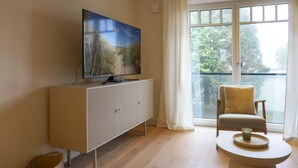 Un salon moderne avec un téléviseur à écran plat posé sur un meuble blanc, une table ronde avec un plateau et une chaise près d’une grande fenêtre donnant sur des arbres.