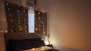 A bed with rose petals and candles, a bedside table with a lamp, and curtains with string lights.